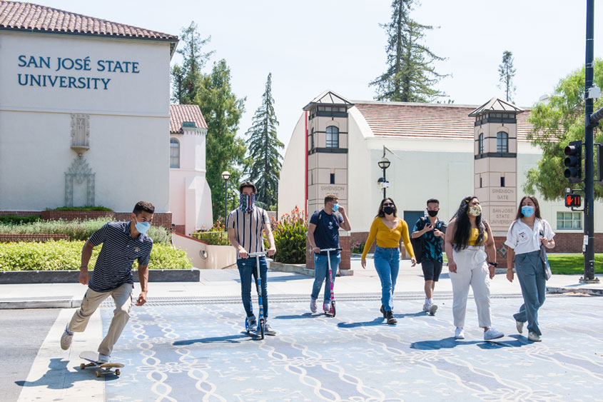 Students crossing the street and one skateboarding with masks on.