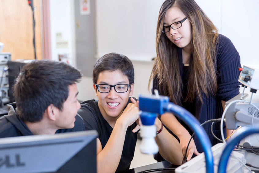 Three engineering students smiling and sitting on a desk with computer equipment.