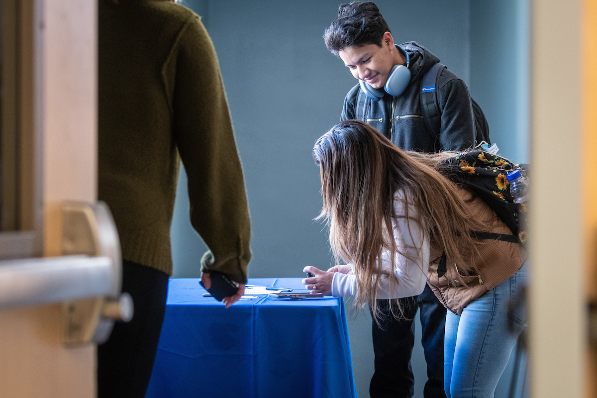 Student leaning down and writing name down at booth, fellow student watches approvingly.