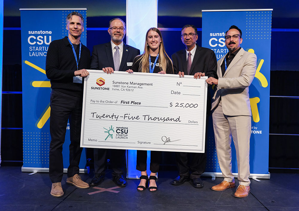 A female, college-age startup founder poses onstage with two sponsors, two hosts, and a giant check for $25,000