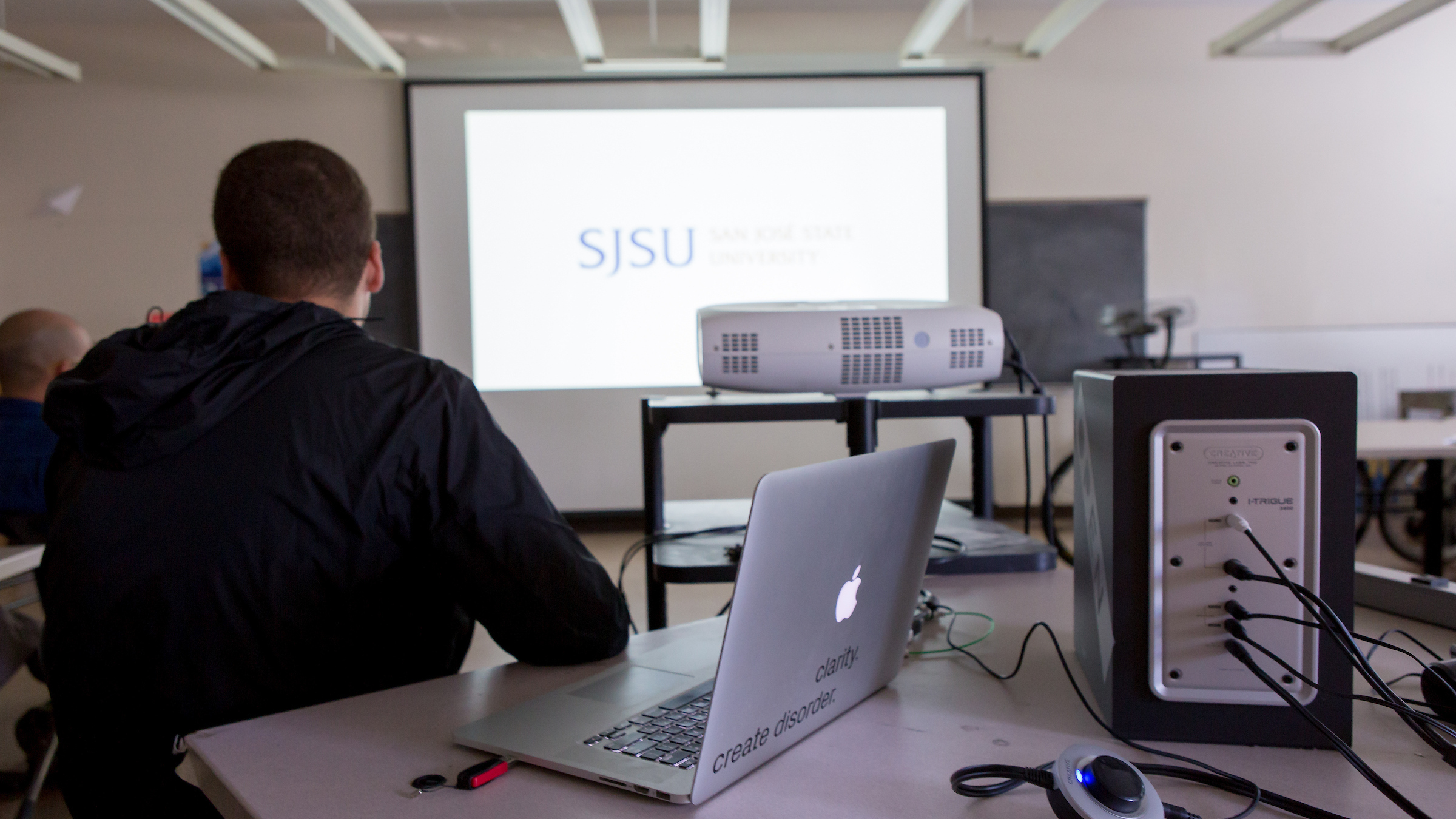 A projector screen in the background shows a gold and blue SJSU logo; in the midground there is a new projector; in the foreground a student is sitting facing the projector screen with a laptop open and there is a box with cables going into it.