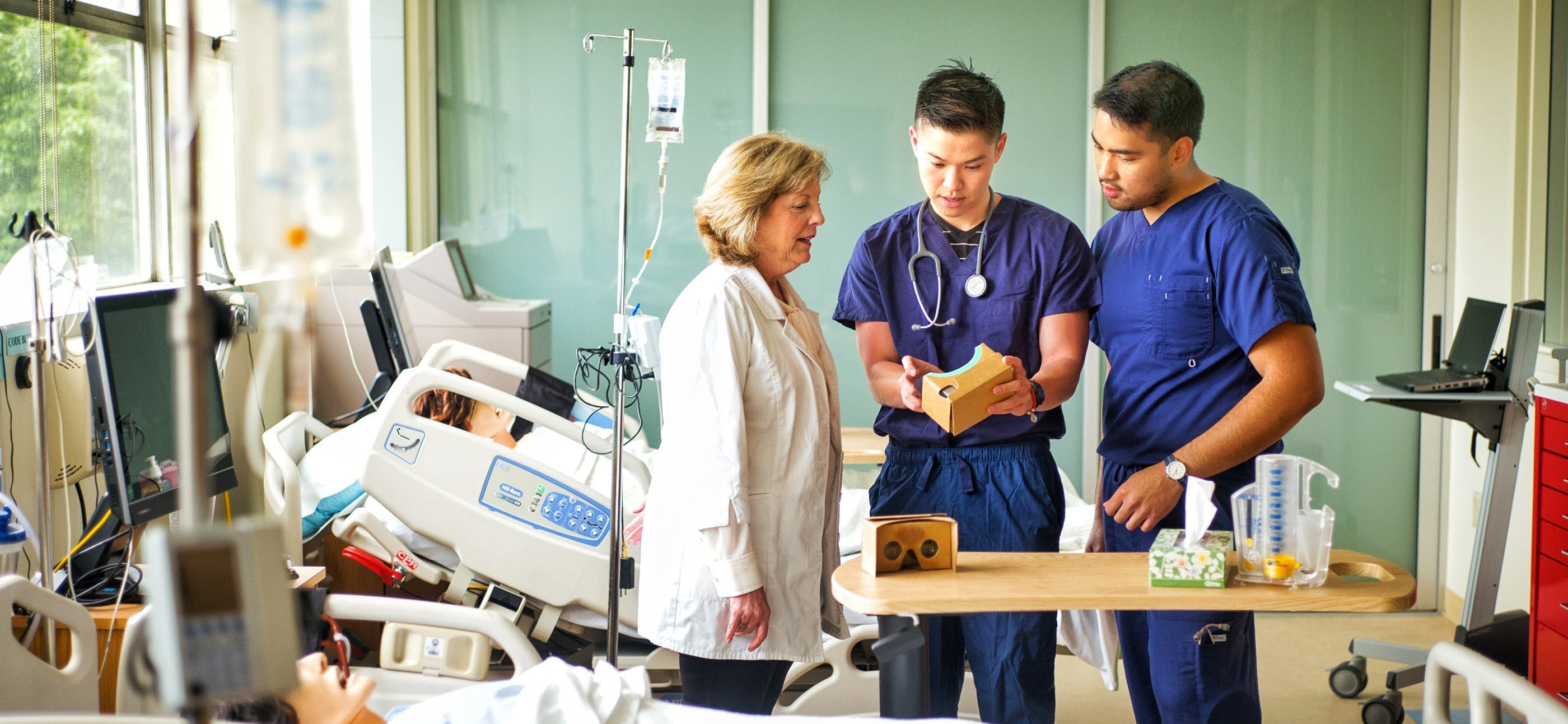 Three nurses, two in blue scrubs and one in a white coat, using virtual reality technology to learn in a nursing simulation lab.