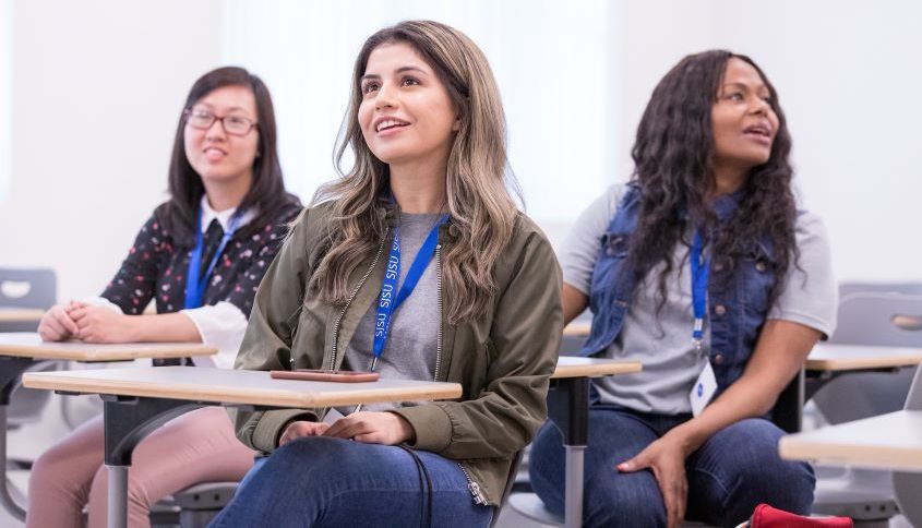 Three students smile while listening at desks in a presentation setting