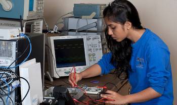 a person works on a circuit board with computers around her