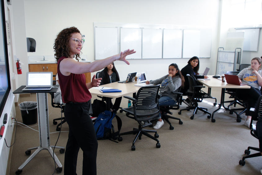 A professor with their arm extended out while talking to the classroom.