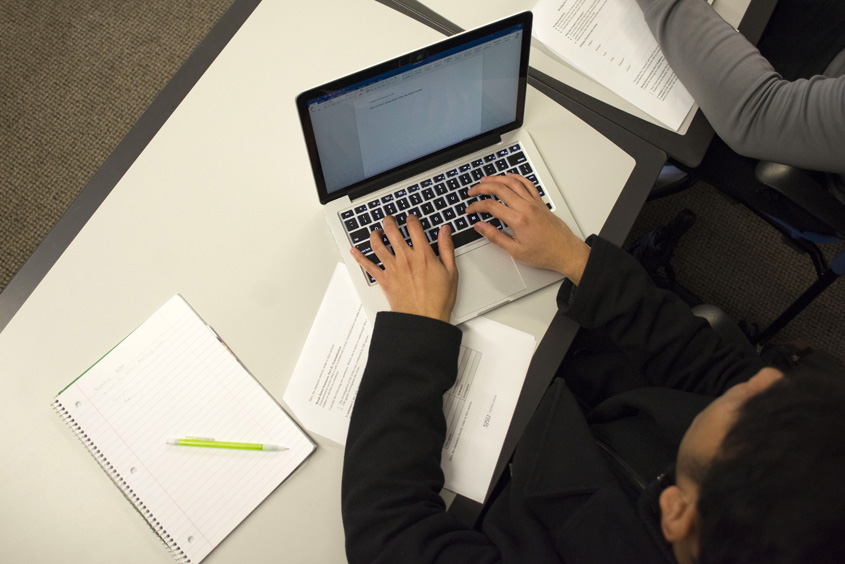 Student at desk typing on laptop with notepad and pen next to them